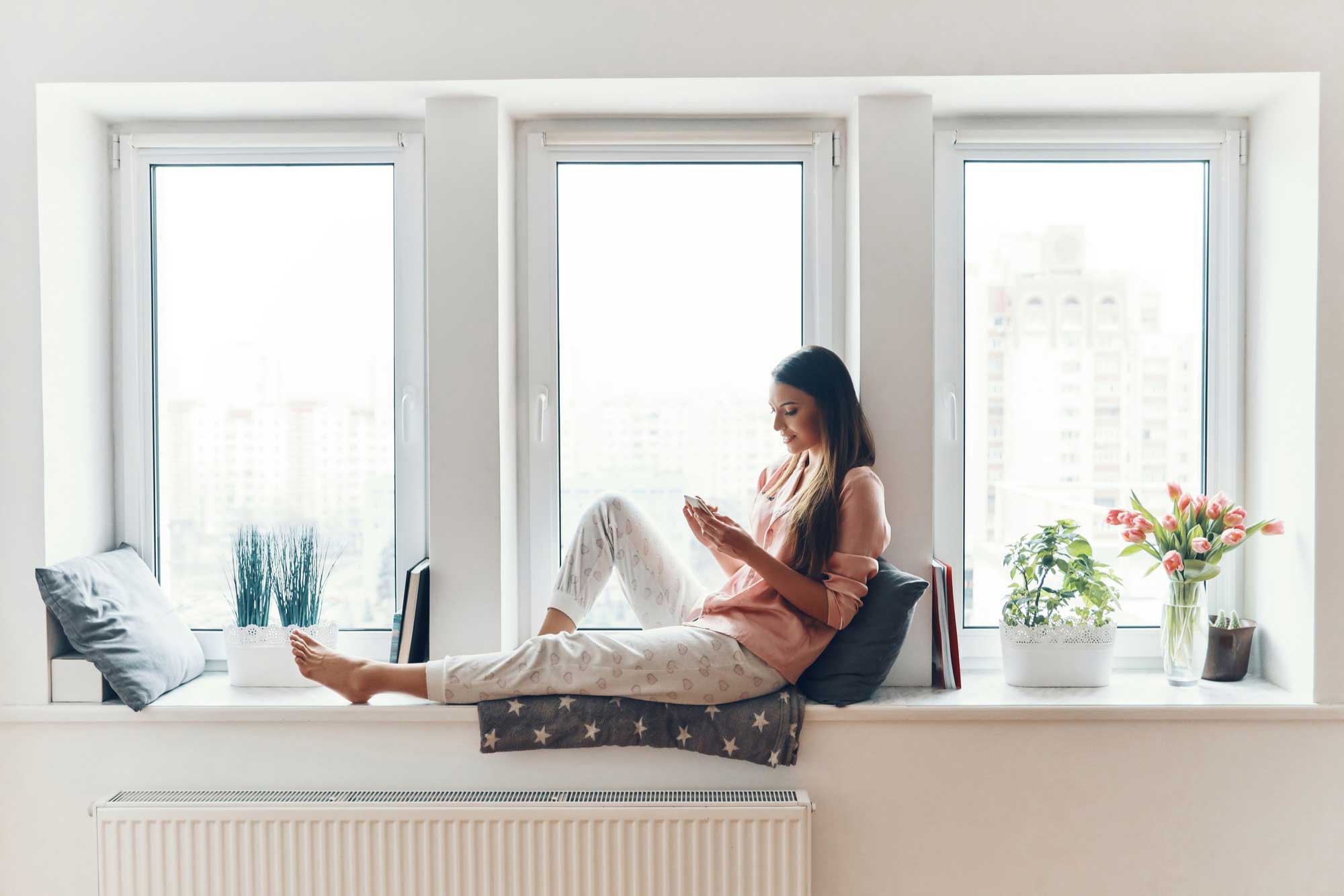 Woman sitting on an indoor window seat using a smartphone beside modern white Casement Windows Atlanta, showcasing bright natural light, clean interior design, and stylish home decor.