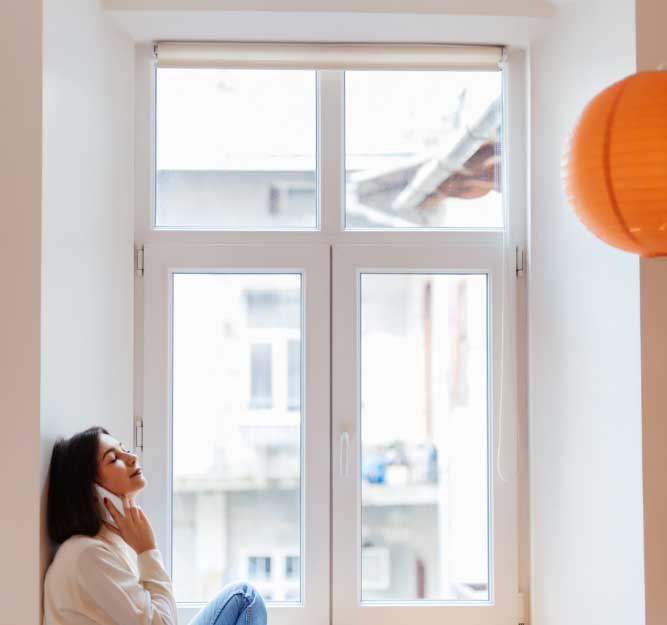 Woman relaxing indoors while talking on a phone beside large white-framed Casement Windows Atlanta, highlighting bright natural light, modern window design, and a comfortable living space.